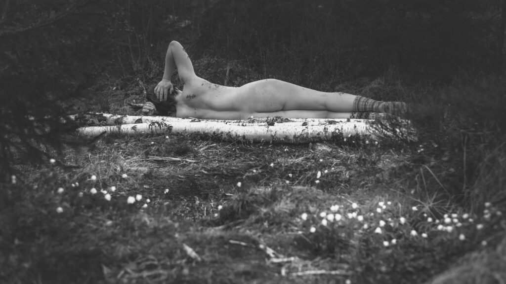 Mustavalkokuvassa on valokuvaaja Minna Alaspää maassa makaavien koivunrunkojen takana, vain villasukat jalassaan.
